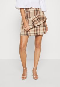 Brązowa mini spódnica w kratę z drapowanym detalem falbany i dopasowanym krojem, wykonana z lekkiego materiału. Połączona z plecionymi białymi obcasami.