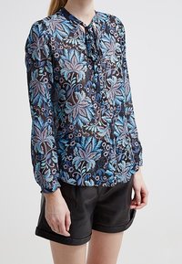 Blommig blus med en mörk bakgrund och blå och lila bladmönster, långa ärmar och knytkrage, kombinerad med svarta shorts.