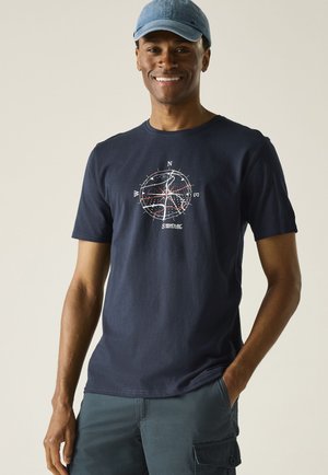 Man wearing a blue cap, navy t-shirt with a compass graphic, and dark cargo shorts, smiling with one hand in pocket.