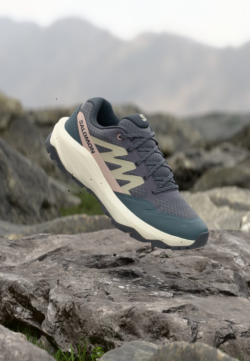 Sötétszürke és bézs Salomon túracipő a levegőben, sziklás terep felett, elmosódott hegyvidéki háttérrel.