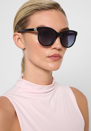 Donna con capelli biondi pettinati all'indietro, che indossa occhiali da sole neri a forma di occhio di gatto e una canotta rosa chiaro su uno sfondo semplice.