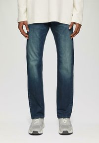 Donkerblauwe denim jeans met een rechte pasvorm, een lichte slijtplek en een zachte textuur; gecombineerd met grijze sportschoenen.