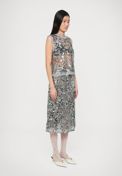 Γκρι διάφανο φόρεμα με διακοσμητικά παγιέτες, εφαρμοστό μπούστο και φούστα μέχρι το γόνατο. Συνδυάζεται με ανοιχτόχρωμα καλσόν από δαντέλα και μυτερά επίπεδα παπούτσια.