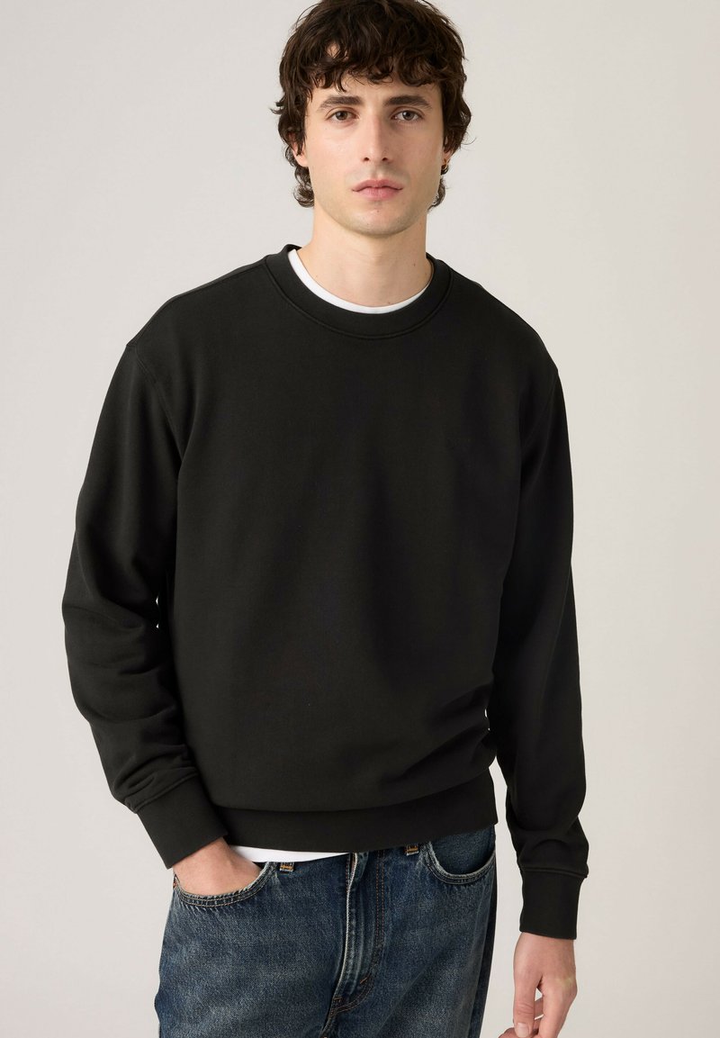 Jeune homme aux cheveux bouclés et foncés portant un sweat-shirt noir à col rond sur une chemise blanche et un jean bleu, debout devant un fond beige uni.