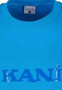 Light blue cotton t-shirt with a round neckline and short sleeves. Features the brand name "KANÍ" in a slightly raised, glossy blue font.