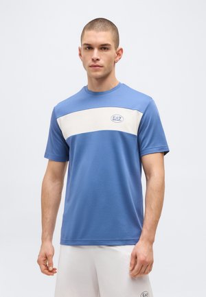 Jeune homme aux cheveux courts portant un t-shirt bleu et blanc EA7 Emporio Armani et un short blanc, se tenant devant un fond uni.