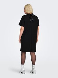 Black short-sleeve dress with a tie bow at the back and a vertical opening. Model wears silver ankle boots and black tights. Smooth fabric.