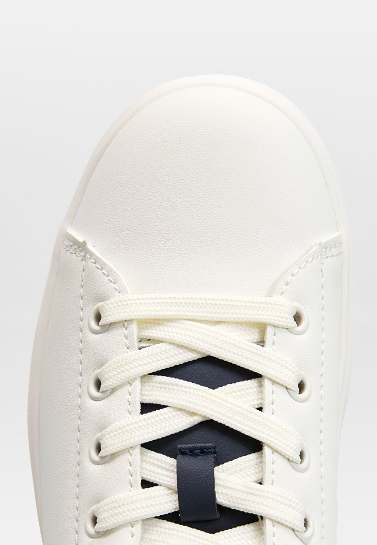 Baskets blanches avec un dessus lisse en synthétique, présentant des accents en maille et des lacets plats. Le design comprend un bout arrondi et une semelle texturée.