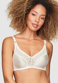 Beige spetstopp med en mjuk, glänsande yta. Har en v-ringning, justerbara axelband och en dekorativ knapp i mitten.