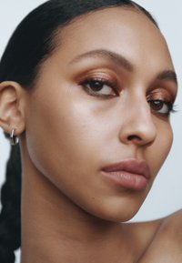 Gros plan sur un visage avec une peau lisse, des yeux marron chaud, un fard à paupières cuivré et une couleur lèvres subtile. Une petite boucle d'oreille en argent est portée.