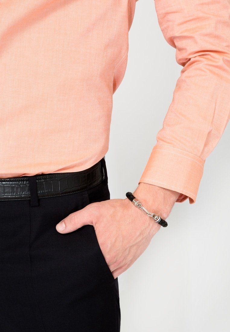 Main droite dans la poche du pantalon noir, portant un bracelet en cuir noir tressé avec une fermeture métallique, associé à une chemise pêche à manches longues et une ceinture noire.
