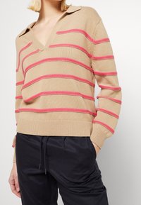 Beżowy sweter w różowe poziome paski, z dekoltem w serek i kołnierzykiem; teksturowana dzianina. Noszony z ciemnymi spodniami z elastycznym pasem.