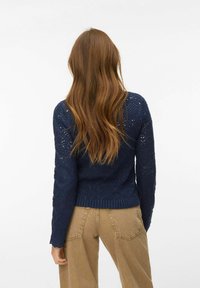 Femme aux longs cheveux bruns portant un pull en maille bleu marine à motifs et un pantalon beige, debout de dos sur un fond blanc.