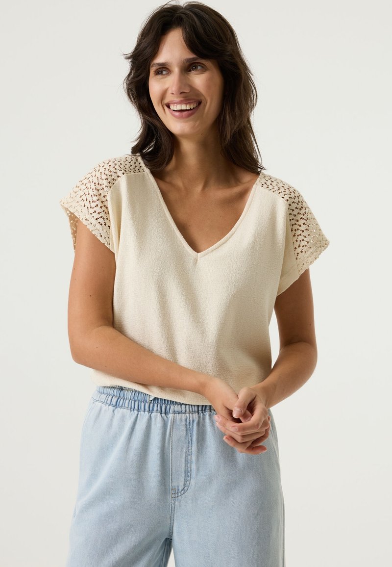 Crèmewitte top met een V-hals, korte mouwen en gehaakte details op de schouders; gecombineerd met lichtblauwe, ontspannen broek.