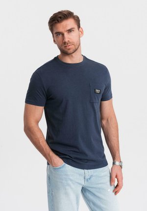 Camiseta de algodón azul marino con cuello redondo, mangas cortas y un bolsillo en el lado izquierdo del pecho que presenta una etiqueta negra. Llevada con jeans de color azul claro.