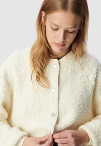 Veste en polaire crème avec une surface texturée, dotée d'une fermeture à boutons et d'un col côtelé. Des mains ajustent la fermeture.