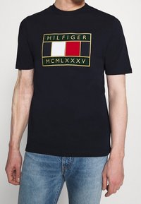 Muž v námořnickém modrém tričku s obdélníkovým logem Tommy Hilfiger, které obsahuje červené, bílé a modré bloky a římské číslice, spárovaném s modrými džínami.