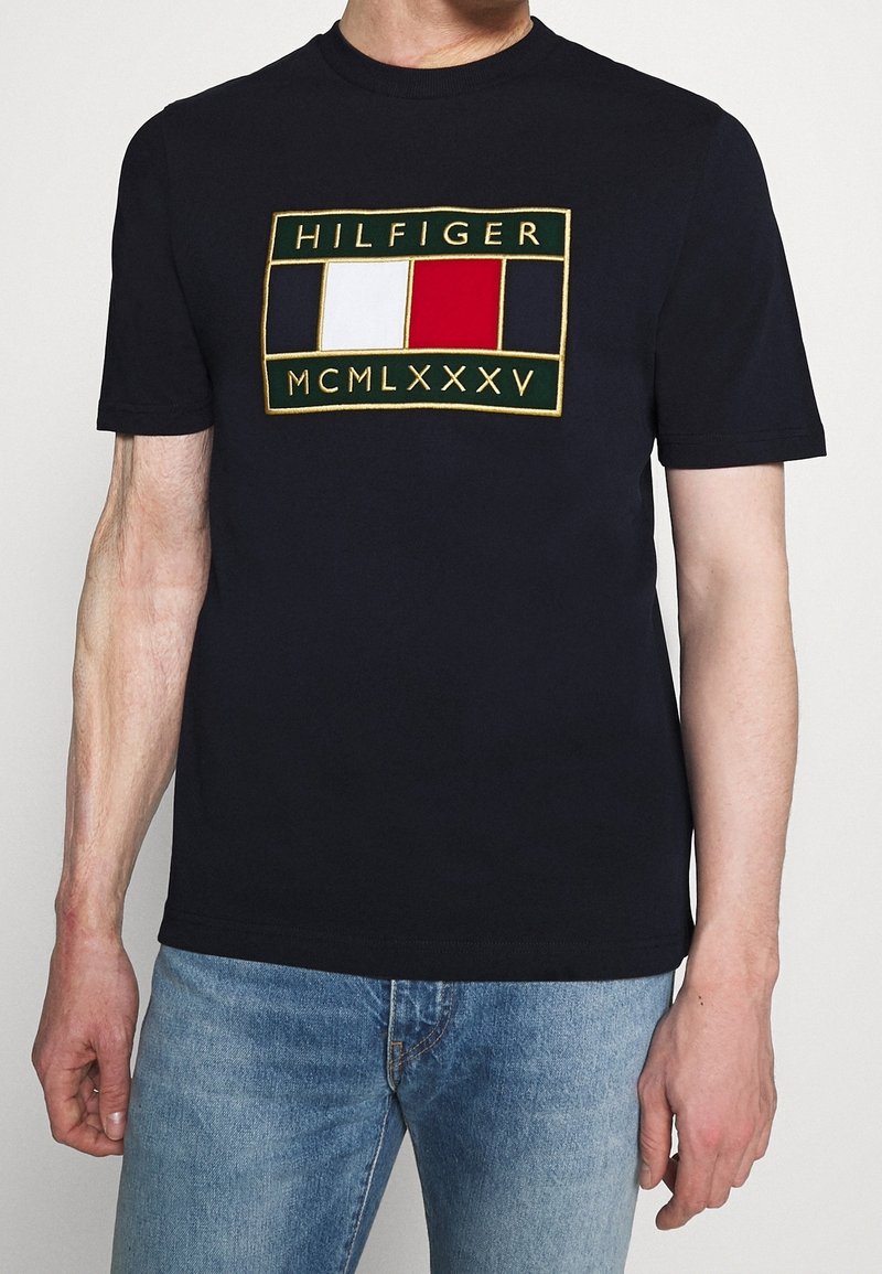 Muž v námořnickém modrém tričku s obdélníkovým logem Tommy Hilfiger, které obsahuje červené, bílé a modré bloky a římské číslice, spárovaném s modrými džínami.