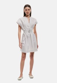 Weißes kurzes Kleid mit kleinem Kragen, plissiertem Rock und Stoffgürtel. Kapuze an den Ärmeln sorgt für einen strukturierten Look. Glatte, leichte Textur.
