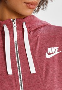 Red cotton hoodie featuring a silver zipper, white drawstrings, and a prominent white Nike logo on the chest. Textured fabric visible.