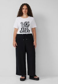 Weißes T-Shirt mit schwarzem grafischen Text "FEEL GREAT" und floralem Design. Lockere schwarze Hose mit Kordelzug, getragen mit schwarzen Plateausandalen.