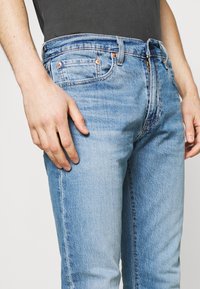 Ljusblå denimjeans med urtvättad textur, femficksdesign, kopparnitar och knäppning med knapp och dragkedja. Modellens hand vilar på låret.