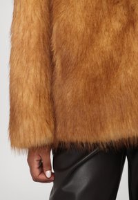 Veste en fausse fourrure marron chaud avec une texture duveteuse, présentant une manche mi-longue et un pantalon en cuir noir lisse.