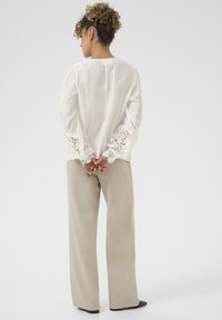 Hvid langærmet bluse med blondekant ved ærmerne, parret med beige vide bukser. Stoffet ser let og tekstureret ud.
