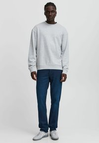 Sudadera gris con puños y dobladillo acanalados, combinada con jeans de denim oscuro y zapatillas blancas, de pie sobre un fondo neutro.