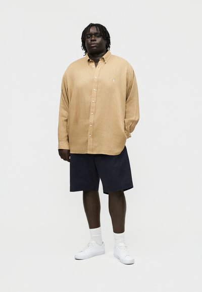 Άνδρας που στέκεται φορώντας μπεζ μακρυμάνικο πουκάμισο με κουμπιά, σκούρες μπλε βερμούδες μέχρι το γόνατο, λευκές κάλτσες και λευκά αθλητικά παπούτσια, πάνω σε απλό φόντο.