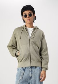 Olivgrüner Reißverschluss-Hoodie mit vorderen Taschen, verstellbarem Kordelzug-Hood und weicher Textur, kombiniert mit hellblauen Denim-Latzhosen.