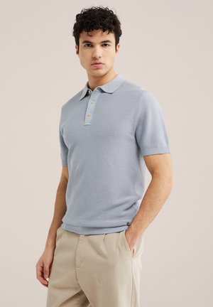 Mladý muž s kučeravými tmavými vlasmi, oblečený v svetlomodré tričko polo s krátkym rukávom a béžové nohavice, stojaci s jednou rukou v vrecku na jednoduchom pozadí.