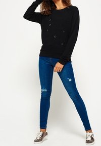 Donna che indossa una felpa nera con nomi di città e jeans skinny strappati blu, in piedi con una mano alzata verso la testa e le gambe divaricate.