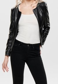 Veste de moto en simili cuir noire avec un design ajusté, fermeture éclair et poches avant, portée sur un débardeur blanc et un jean noir.