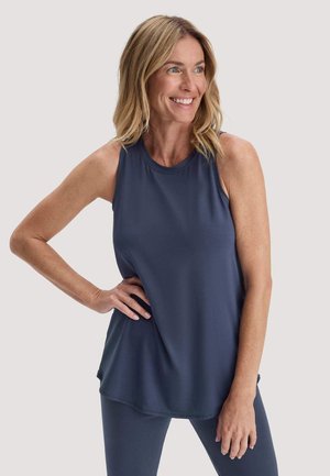 Lächelnde Frau mit blonden Haaren, die ein ärmelloses marineblaues Oberteil und passende Leggings trägt, und mit einer Hand in der Hüfte posiert.