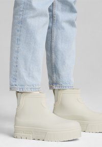 Bottines en caoutchouc blanches à la semelle épaisse et aux détails texturés, associées à un jean en denim bleu clair, affichant un design épuré et moderne.