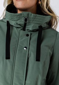 Groene jas met een hoge kraag, zwarte trekkoorden, drukknoopsluiting en een rits met glanzende accenten. Gladde stoftextuur.