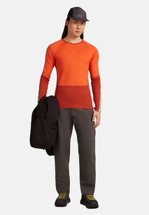Persoon in een oranje en rode lange mouwen shirt, donkere broek, zwarte pet, die een zwarte jas vasthoudt en tegen een witte achtergrond staat.