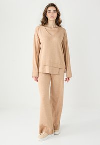 Top beige a maniche lunghe con scollo a V, design a strati, abbinato a pantaloni a gamba larga coordinati, realizzati in un morbido tessuto testurizzato.