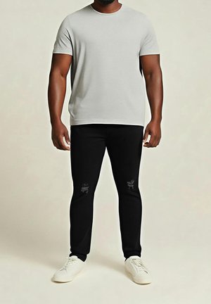 Hombre con camisa de manga corta gris claro, pantalones negros con discretas rodilleras y zapatillas blancas, de pie frente a un fondo beige liso.