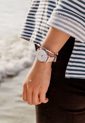 Montre en argent avec un cadran blanc et un bracelet en cuir beige, portée au poignet à côté d'un bracelet métallique fin. Manches en tissu rayé visibles.
