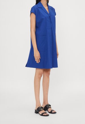 Femme portant une robe bleu royal à manches courtes, longueur genou, avec un col et des poches avant, associée à des sandales à talons ouverts noires.