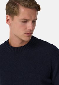 Tmavě modrý pletený svetr s žebrovaným kulatým výstřihem a hladkou strukturou, zobrazený na modelce s krátkými vlasy a neutrálním výrazem.