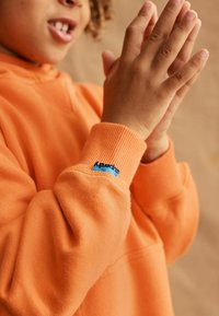 Felpa arancione con polsini a costine, caratterizzata da un piccolo logo blu sulla manica. Tessuto morbido e testurizzato con una vestibilità rilassata.
