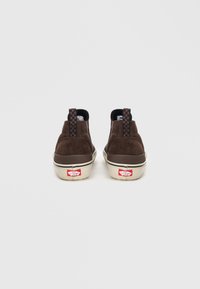 Brune semskede slip-on sko med sjakkmønster stoff hælfliker og beige gummisåler, med en rød logo bak.
