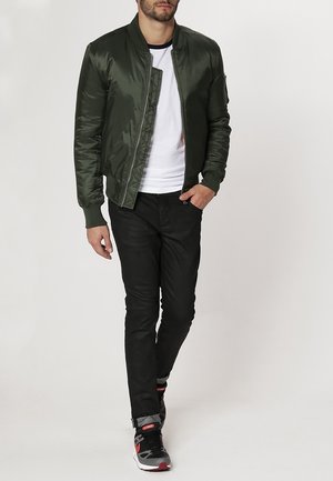 Olive green bomber jacket with a smooth, shiny finish, ribbed collar, and cuffs. Worn over a white T-shirt and paired with black pants and sneakers.