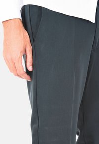 Pantaloni grigi scuri realizzati in tessuto liscio con tasche laterali, caratterizzati da una vestibilità slim e una trama sottile, mostrati con una mano appoggiata di lato.