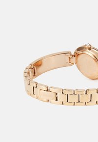 Goldfarbene Uhr mit einem Metallarmband-Design, das polierte Glieder und ein rundes Gehäuse mit einem Kristallakzent aufweist. Graviertes Logo am Band.