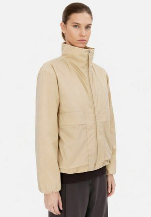 Kvinde iført en beige højhalset dunjakke med lange ærmer og forlommer, kombineret med sorte bukser mod en hvid baggrund.