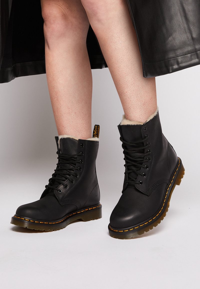 Jambes portant des bottes en cuir noir à lacets avec doublure en fourrure et couture jaune, assorties à une jupe en cuir noir.